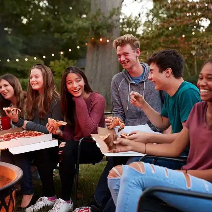 Group of teens laughing and eating pizza near a campfire