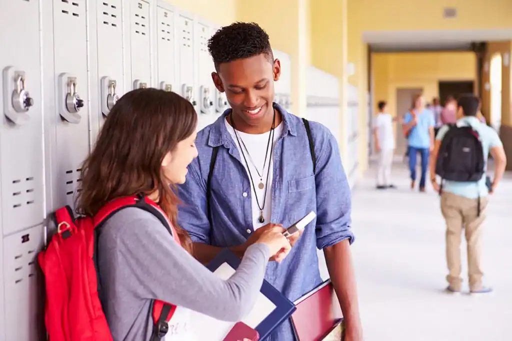Two students in a school hallway smiling while looking at a smartphone