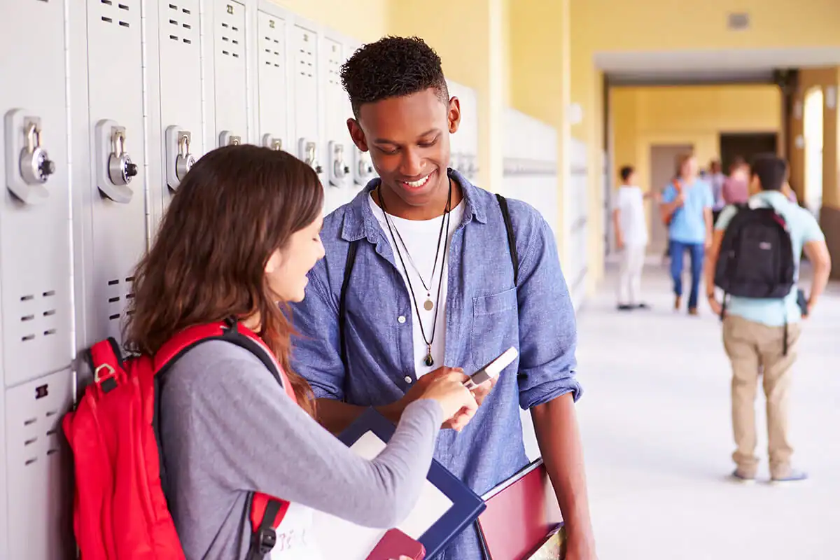 Two students in a school hallway smiling while looking at a smartphone
