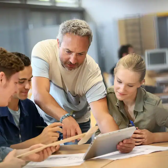 Educator holding a tablet device working alongside three students in a classroom